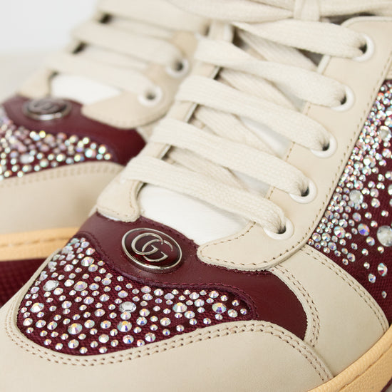 Close-up of a luxury sneaker featuring cream leather panels, deep burgundy leather overlays, and extensive rhinestone embellishments. A silver-tone interlocking GG logo is visible on the side, identifying the brand as Gucci.