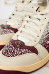 Close-up of a luxury sneaker featuring cream leather panels, deep burgundy leather overlays, and extensive rhinestone embellishments. A silver-tone interlocking GG logo is visible on the side, identifying the brand as Gucci.