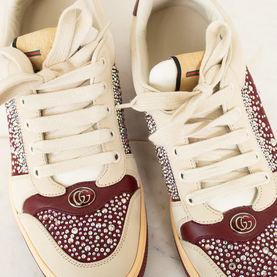 A pair of Gucci low-top sneakers in cream and burgundy, embellished with rhinestones and featuring the iconic interlocking GG logo on the toe box, displayed on a light marble surface.