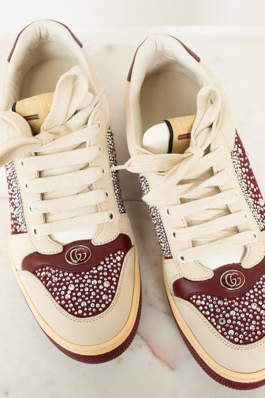 A pair of Gucci low-top sneakers in cream and burgundy, embellished with rhinestones and featuring the iconic interlocking GG logo on the toe box, displayed on a light marble surface.