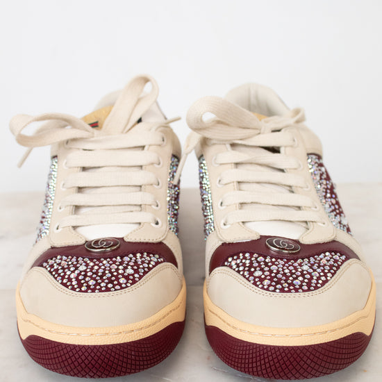 A pair of low-top sneakers with cream-colored leather uppers, burgundy rubber soles, and sparkling rhinestone embellishments on the sides and toe caps, displayed on a light marble surface against a white background.
