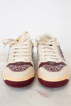 A pair of low-top sneakers with cream-colored leather uppers, burgundy rubber soles, and sparkling rhinestone embellishments on the sides and toe caps, displayed on a light marble surface against a white background.