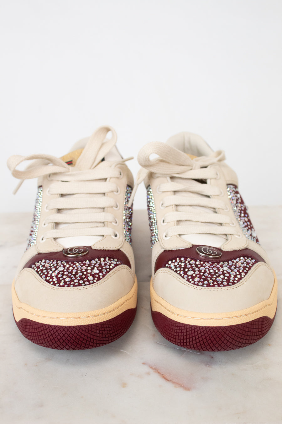 A pair of low-top sneakers with cream-colored leather uppers, burgundy rubber soles, and sparkling rhinestone embellishments on the sides and toe caps, displayed on a light marble surface against a white background.