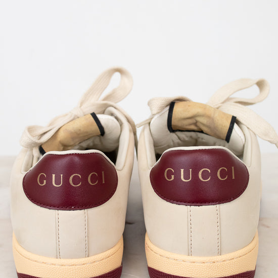 A pair of Gucci low-top sneakers shown from the back, featuring cream leather uppers, burgundy leather heel tabs with gold 'GUCCI' lettering, and cream midsoles with burgundy outsoles.