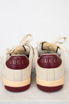 A pair of Gucci low-top sneakers shown from the back, featuring cream leather uppers, burgundy leather heel tabs with gold 'GUCCI' lettering, and cream midsoles with burgundy outsoles.