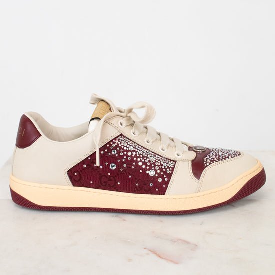 A low-top sneaker with cream-colored panels, burgundy overlays, and a sparkling rhinestone-embellished side panel, resting on a white marble surface against a white background.
