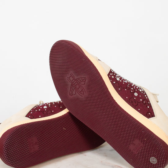 A pair of women's low-top sneakers shown from the sole side, featuring deep burgundy textured rubber soles with a debossed logo and rhinestone-embellished burgundy overlays on a cream-colored upper.