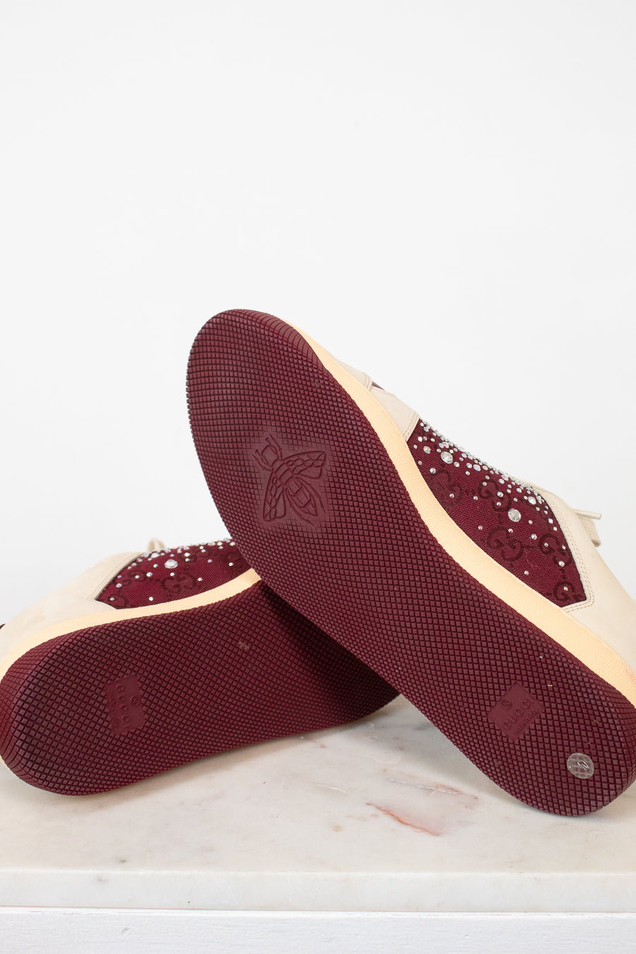 A pair of women's low-top sneakers shown from the sole side, featuring deep burgundy textured rubber soles with a debossed logo and rhinestone-embellished burgundy overlays on a cream-colored upper.