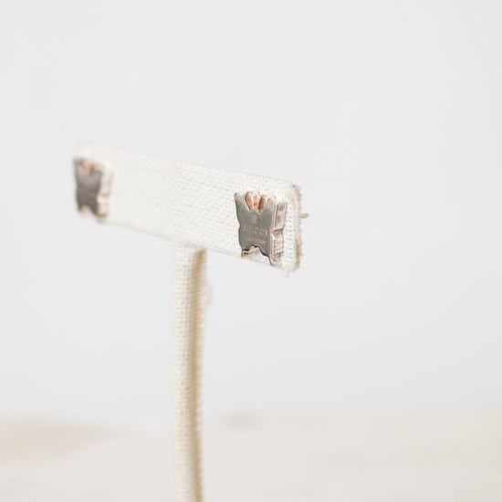 A side profile view of a silver butterfly studs from GUCCI  resting on a beige earring stand.
