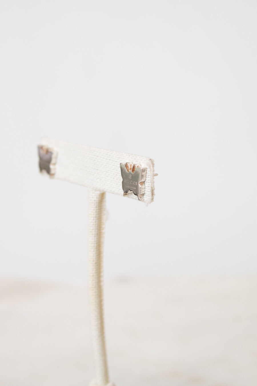 A side profile view of a silver butterfly studs from GUCCI  resting on a beige earring stand.