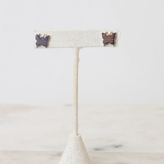 Pair of silver-tone GUCCI butterfly-shaped stud earrings with engraved logo detailing. Resting on a beige earring stand on a marble and white background.