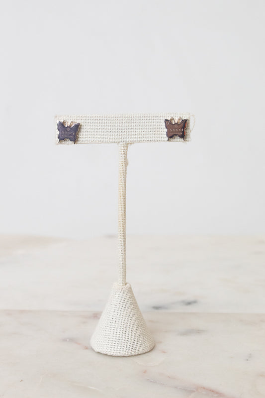 Pair of silver-tone GUCCI butterfly-shaped stud earrings with engraved logo detailing. Resting on a beige earring stand on a marble and white background.