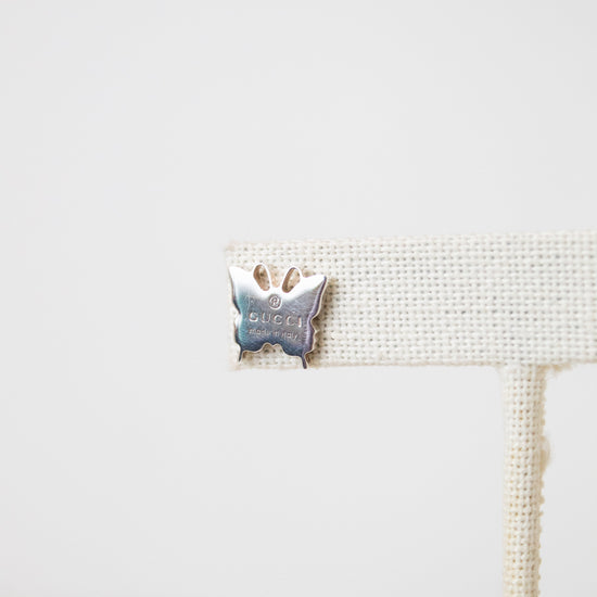 A close-up butterfly shaped stud on a beige earring stand, stating with the word GUCCI engraved. 