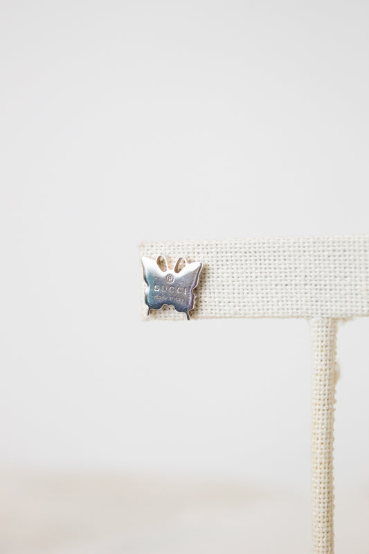 A close-up butterfly shaped stud on a beige earring stand, stating with the word GUCCI engraved. 