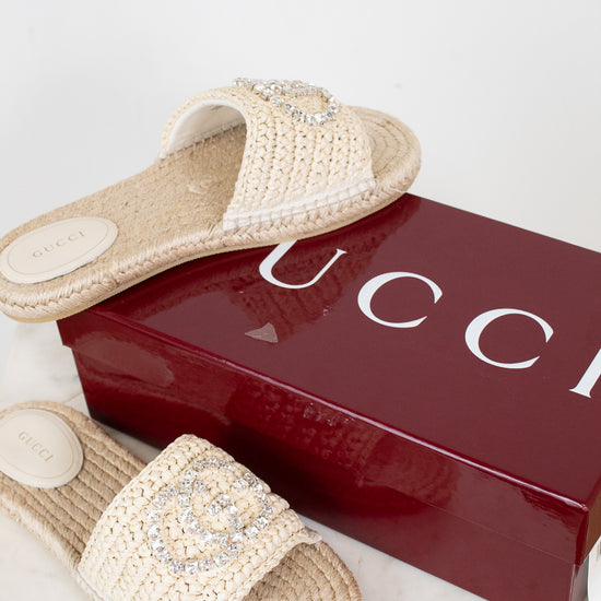 A pair of Gucci woven straw slide sandals with crystal-embellished straps, displayed next to a glossy dark red Gucci shoebox on a light marble surface.