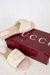 A pair of Gucci woven straw slide sandals with crystal-embellished straps, displayed next to a glossy dark red Gucci shoebox on a light marble surface.