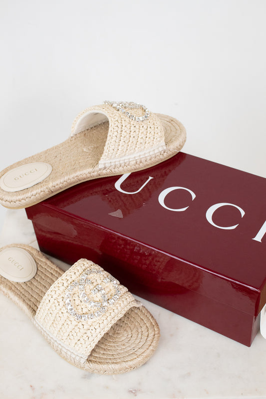 A pair of Gucci woven straw slide sandals with crystal-embellished straps, displayed next to a glossy dark red Gucci shoebox on a light marble surface.