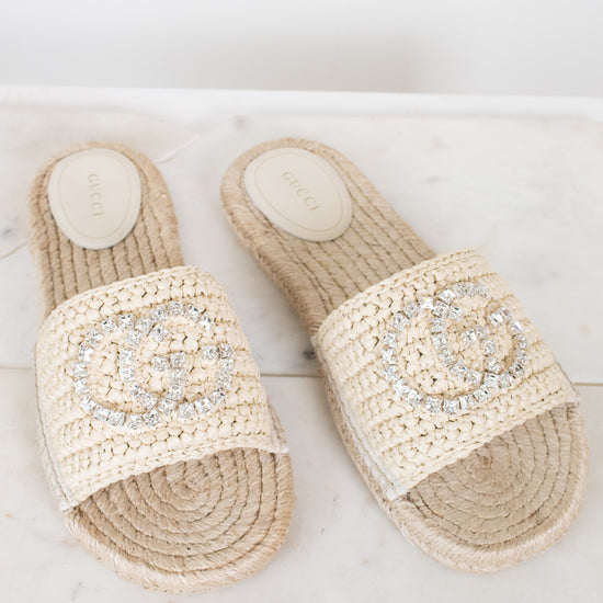 A pair of Gucci woven espadrille slide sandals in beige raffia, featuring a prominent crystal-embellished interlocking GG logo on the strap and a visible Gucci brand insole.