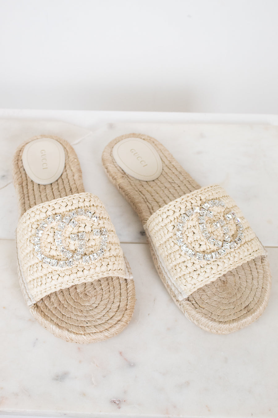 A pair of Gucci woven espadrille slide sandals in beige raffia, featuring a prominent crystal-embellished interlocking GG logo on the strap and a visible Gucci brand insole.