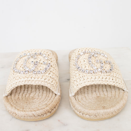 A pair of beige woven slide sandals with a large rhinestone-embellished interlocking GG logo on the top, displayed on a white marble surface against a white background.