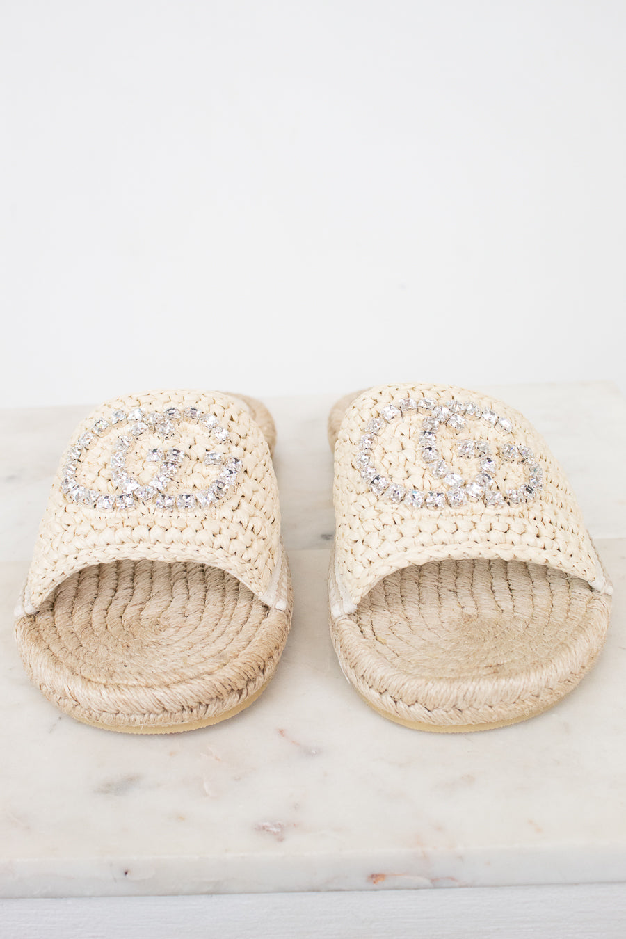 A pair of beige woven slide sandals with a large rhinestone-embellished interlocking GG logo on the top, displayed on a white marble surface against a white background.