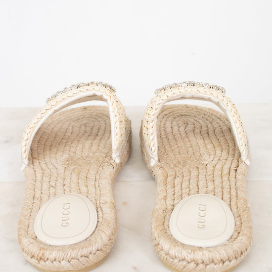 A pair of Gucci espadrille-style slide sandals with woven beige jute soles and white straps embellished with small rhinestones, shown on a light marble surface against a white background.