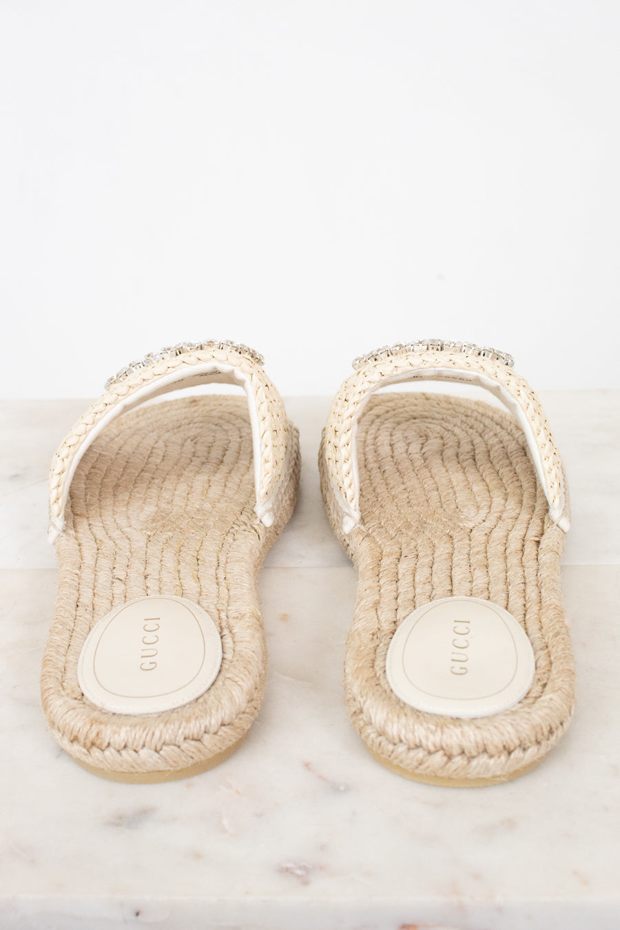 A pair of Gucci espadrille-style slide sandals with woven beige jute soles and white straps embellished with small rhinestones, shown on a light marble surface against a white background.