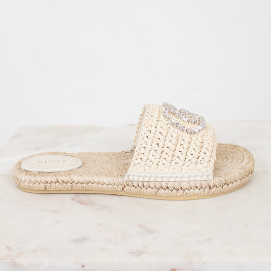 A single beige crochet espadrille-style slide sandal with a rhinestone-embellished strap, resting on a white marble slab against a white background.