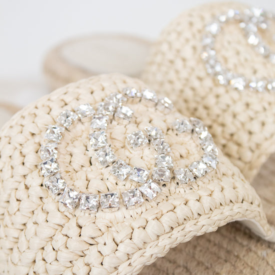 Beige woven straw sandals featuring a large silver rhinestone double-G style logo on the strap, shown in a close-up shot.