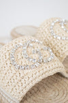 Beige woven straw sandals featuring a large silver rhinestone double-G style logo on the strap, shown in a close-up shot.