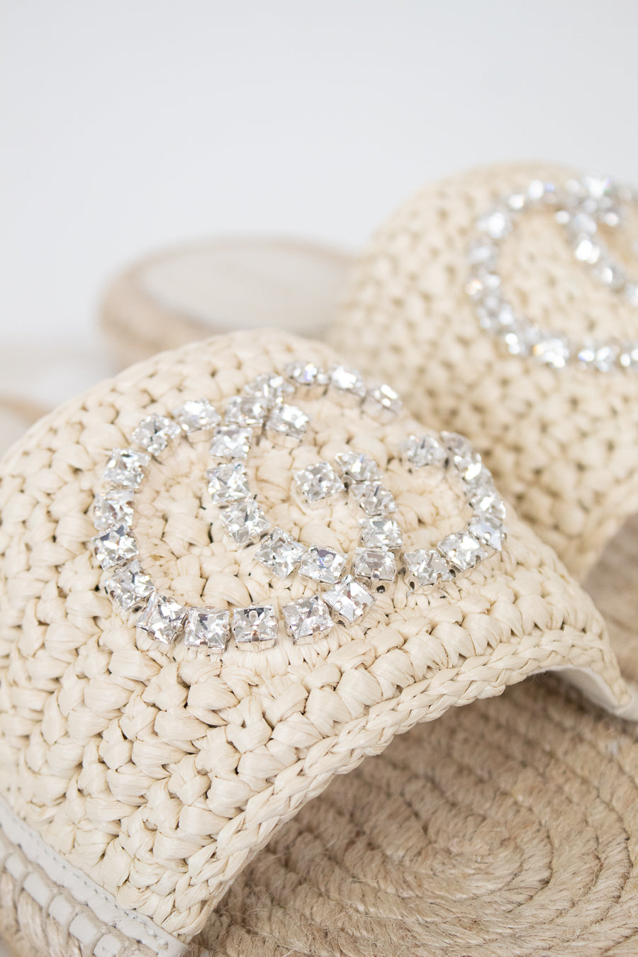 Beige woven straw sandals featuring a large silver rhinestone double-G style logo on the strap, shown in a close-up shot.