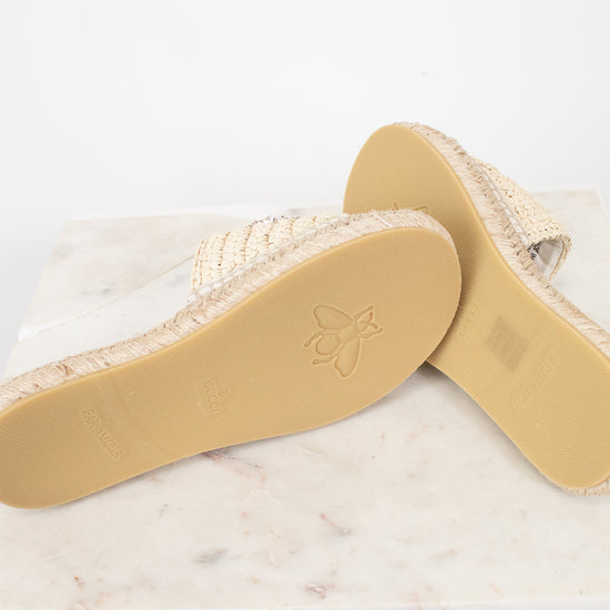 A pair of woven espadrille-style slide sandals shown from the sole side, resting on a white marble slab against a white background.