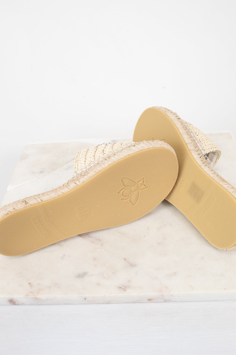A pair of woven espadrille-style slide sandals shown from the sole side, resting on a white marble slab against a white background.