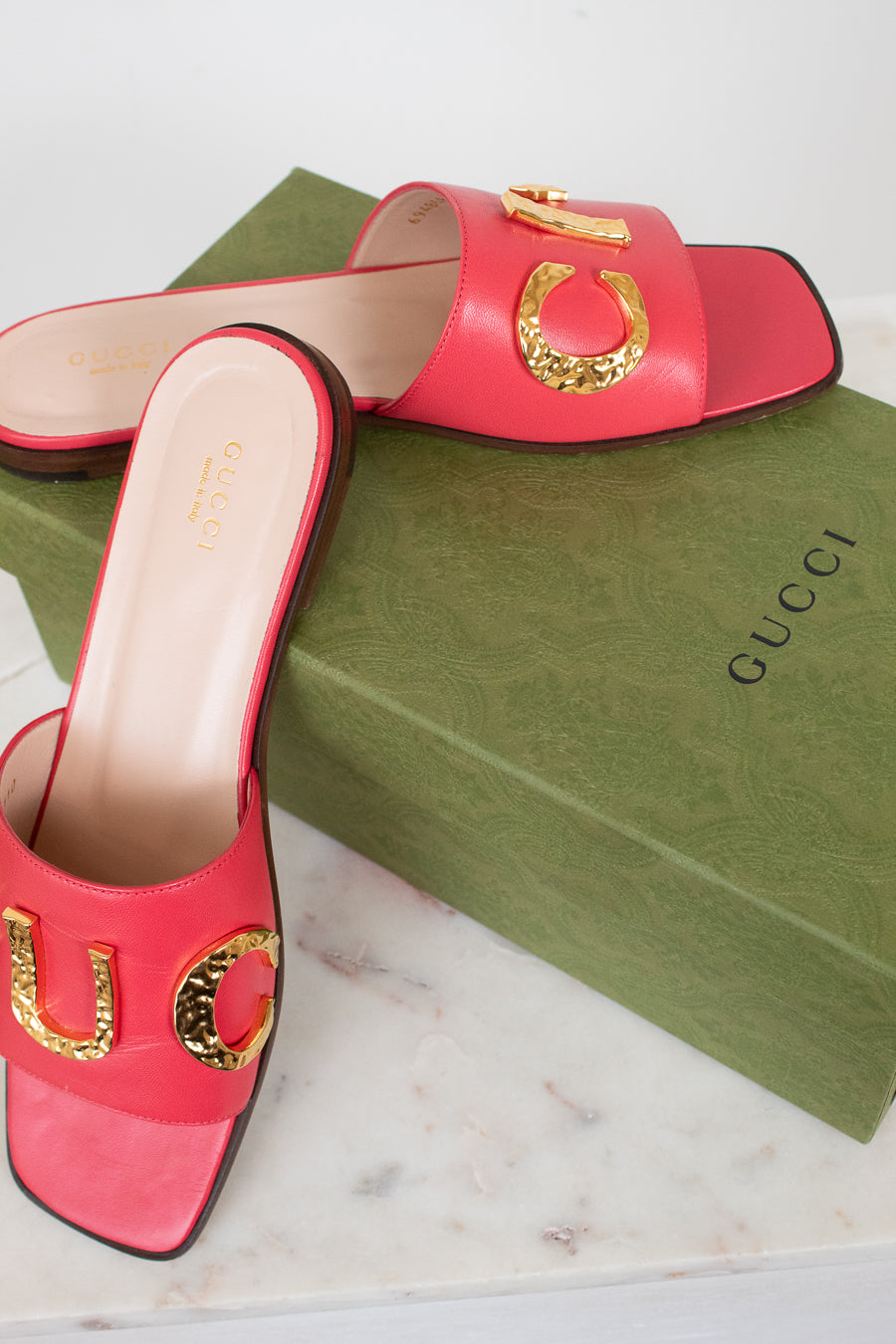 A pair of Gucci pink leather slide sandals with gold-tone horsebit hardware, displayed on top of a green Gucci shoebox on a white marble surface.