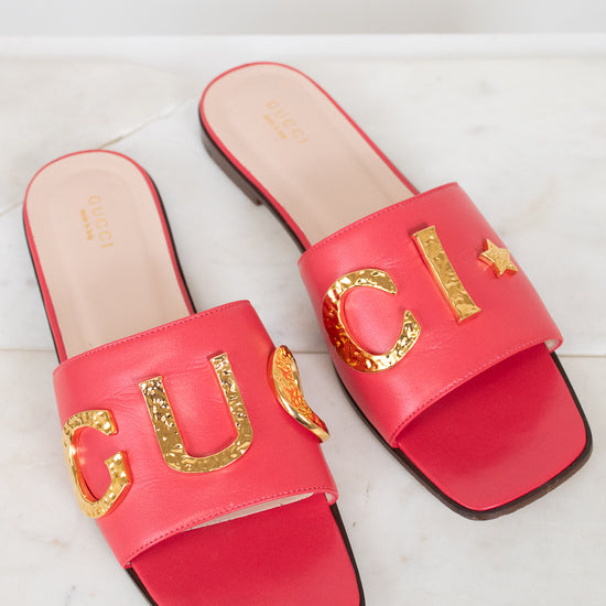 A pair of Gucci red slide sandals with gold metallic 'GUCCI' lettering across the straps, displayed on a light marble surface.