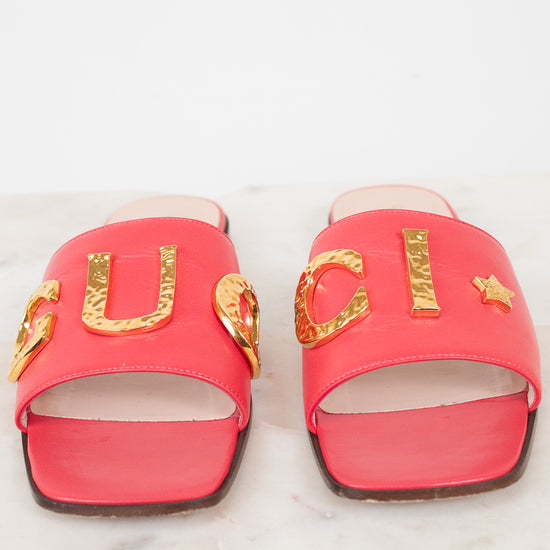 A pair of bright pink Gucci slide sandals with gold-tone metal 'GUCCI' lettering and star details, displayed on a white marble surface against a white background.