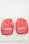 A pair of bright pink Gucci slide sandals with gold-tone metal 'GUCCI' lettering and star details, displayed on a white marble surface against a white background.