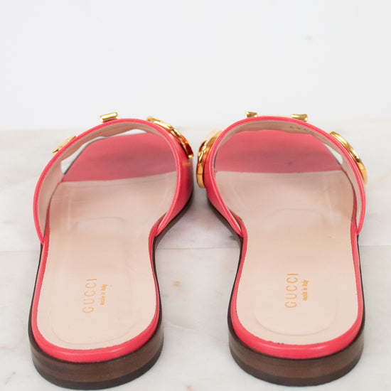 A pair of pink leather Gucci flat slide sandals, with gold-tone letter detailing across the strap, and a dark brown sole, shown from the back on a light marble surface.