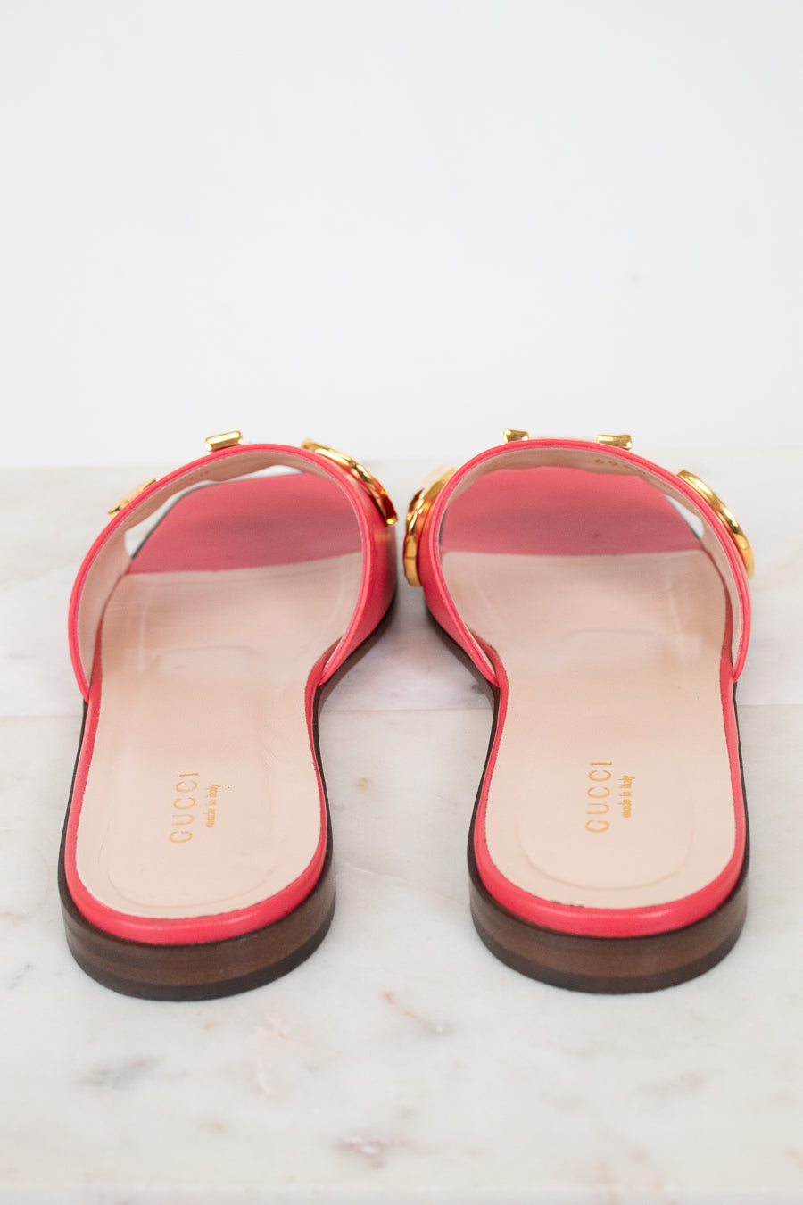 A pair of pink leather Gucci flat slide sandals, with gold-tone letter detailing across the strap, and a dark brown sole, shown from the back on a light marble surface.