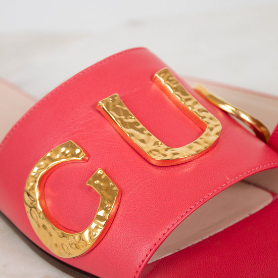 A close-up of a bright pink leather slide sandal featuring large, hammered gold metallic letters across the strap, set against a light marble background.