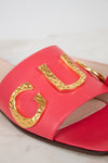 A close-up of a bright pink leather slide sandal featuring large, hammered gold metallic letters across the strap, set against a light marble background.