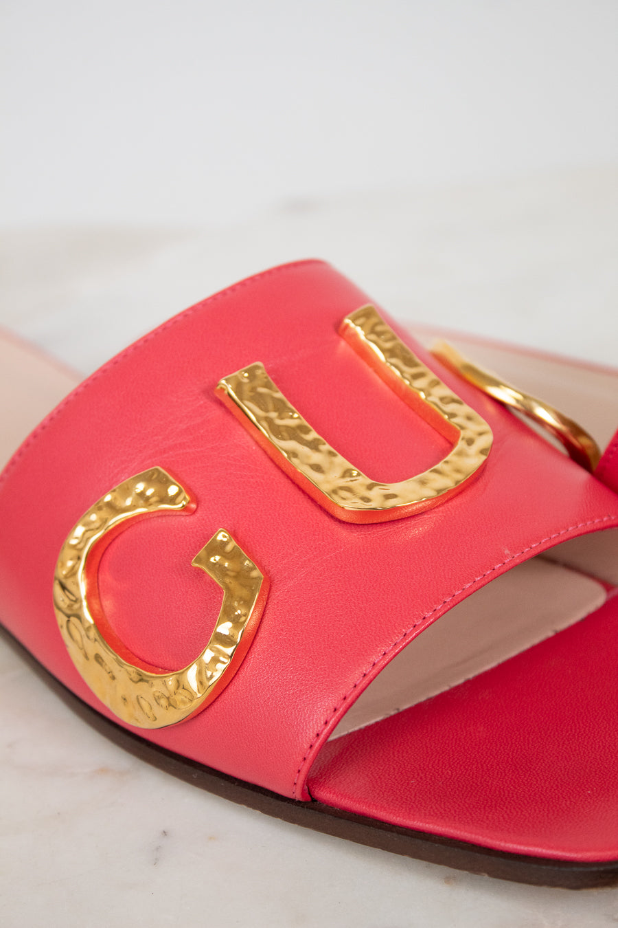 A close-up of a bright pink leather slide sandal featuring large, hammered gold metallic letters across the strap, set against a light marble background.