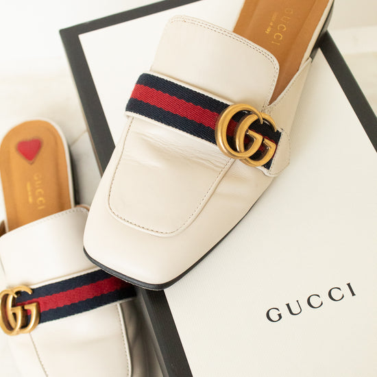 A pair of Gucci white leather mule loafers with the signature red and navy green web stripe and gold double G logo, displayed on top of a white Gucci shoebox.