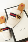 A pair of Gucci white leather mule loafers with the signature red and navy green web stripe and gold double G logo, displayed on top of a white Gucci shoebox.