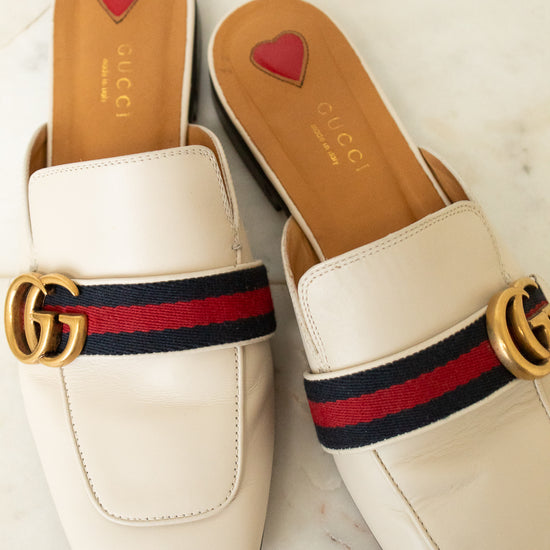 A pair of Gucci square-toe mule loafers in off-white leather, featuring the signature red and navy green web stripe with a gold interlocking GG logo across the top. The insoles are tan with a small red heart logo and the brand name 'GUCCI' printed in gold.