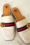 A pair of Gucci square-toe mule loafers in off-white leather, featuring the signature red and navy green web stripe with a gold interlocking GG logo across the top. The insoles are tan with a small red heart logo and the brand name 'GUCCI' printed in gold.