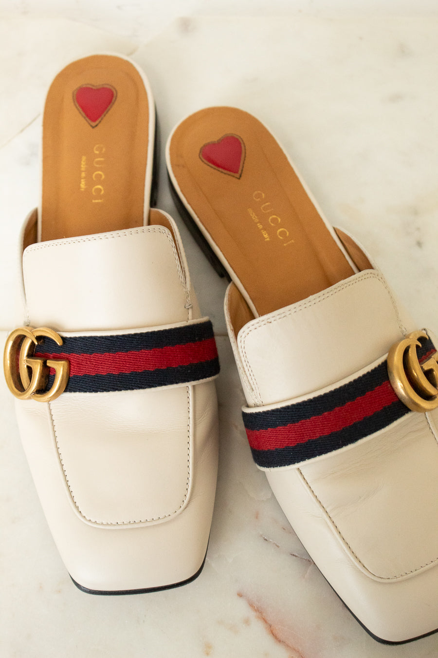 A pair of Gucci square-toe mule loafers in off-white leather, featuring the signature red and navy green web stripe with a gold interlocking GG logo across the top. The insoles are tan with a small red heart logo and the brand name 'GUCCI' printed in gold.