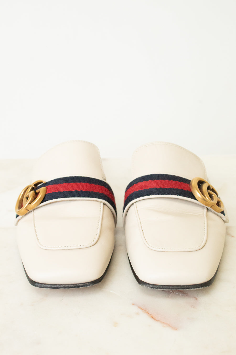 A pair of Gucci square-toe leather loafers in off-white, featuring the signature red and navy green web stripe across the top with a gold-tone interlocking GG logo on each side.