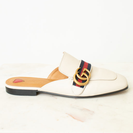 A single Gucci flat mule sandal in off-white leather, featuring the signature red, green, and navy web stripe across the vamp with a gold interlocking GG logo, set against a clean white background.
