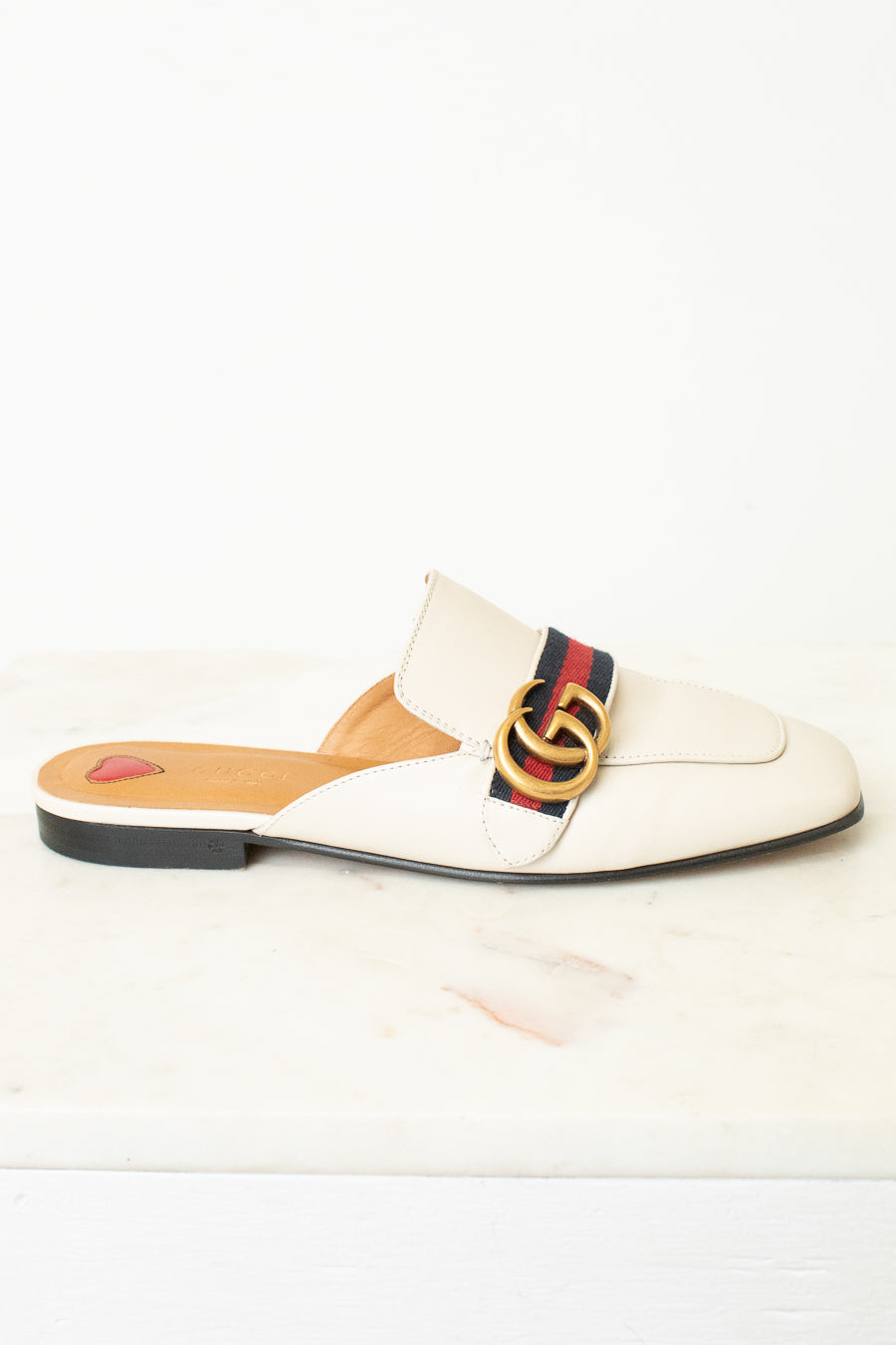 A single Gucci flat mule sandal in off-white leather, featuring the signature red, green, and navy web stripe across the vamp with a gold interlocking GG logo, set against a clean white background.